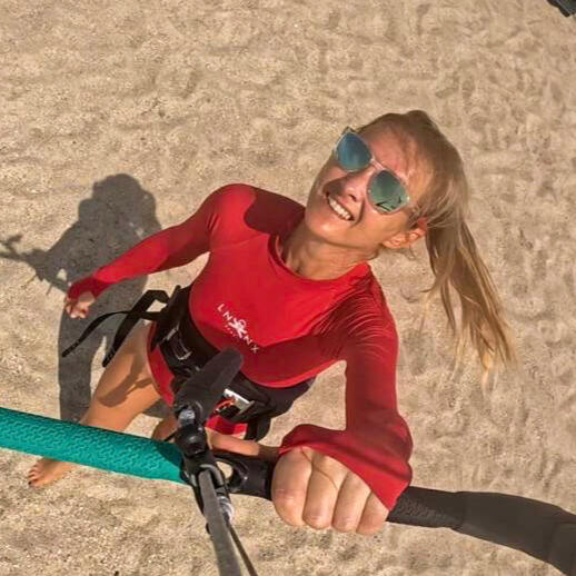 Launching a kite An overhead, point-of-view shot from the perspective of a kitesurfer holding a control bar with teal and black grips. Below, a smiling woman in a long-sleeved red rash guard and a kitesurfing harness stands on a sandy beach, looking up toward the camera. H
